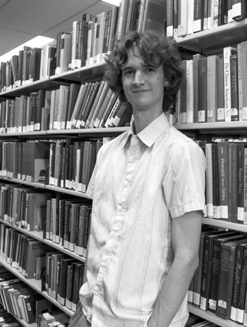 Black and white photograph of Australian cartoonist Stuart McMillen. Standing in front of a book shelf in a library.