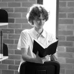 Stuart McMillen in a library, sitting backwards on a chair, reading a book - black and white, wide crop.