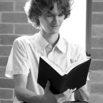 Stuart McMillen in a library, sitting backwards on a chair, reading a book - black and white, mid crop.