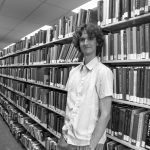 Stuart McMillen standing between library shelves - black and white, wide shot.
