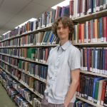 Stuart McMillen standing between library shelves - colour, wide shot.