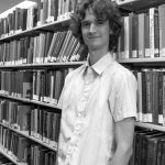 Stuart McMillen standing between library shelves - black and white, mid shot.