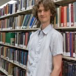 Stuart McMillen standing between library shelves - colour, mid shot.