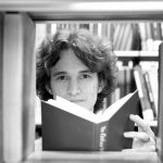 Stuart McMillen in a library, looking between books on a book shelf, holding a book in his hands. Black and white.