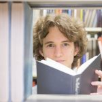 Stuart McMillen in a library, looking between books on a book shelf, holding a book in his hands. Colour.