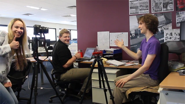 Interns helping with video shoot in coworking office. Standing behind a DSLR video camera tripod on film set.