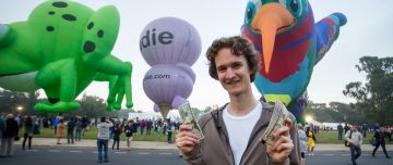 Stuart McMillen at Canberra Balloon Spectacular, holding banknotes in front of colourful hot air balloons