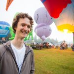 Cartoonist Stuart McMillen in 2018, standing in front of colourful hot air balloons, smiling at camera.