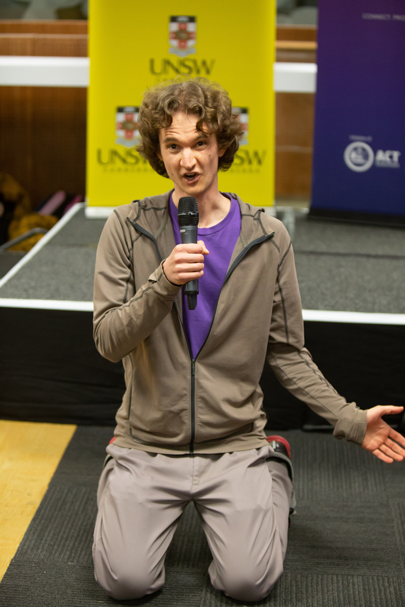 Stuart McMillen public speaking, holding microphone, on the floor dropped to his knees