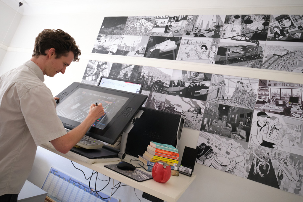 Cartoonist in art studio, drawing at graphics tablet. Rear view of the cartoonist at work at Wacom tablet, with wall of artworks on in background of scene. Stuart McMillen at Gorman Arts Centre